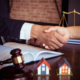 Two people shaking hands over legal documents with a gavel, miniature houses, and scales of justice on a desk.