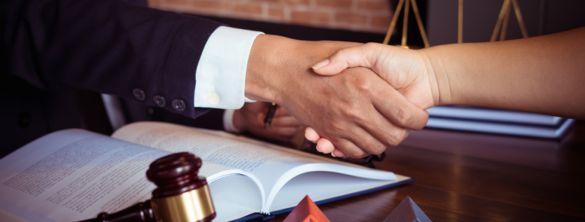 Two people shaking hands over legal documents with a gavel, miniature houses, and scales of justice on a desk.