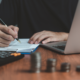 Person signing documents with a pen, calculator, and stacks of coins on a desk, symbolizing financial planning and trust funding.